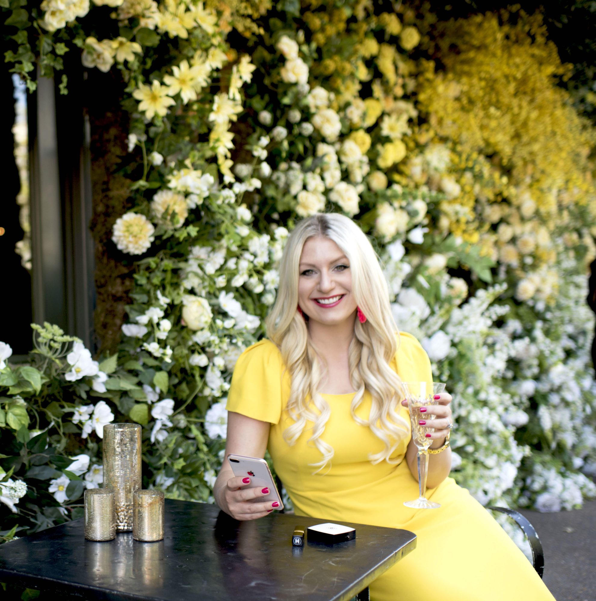 Emily Williams sitting in a yellow dress, holding a glass of champagne, in front of a flower wall